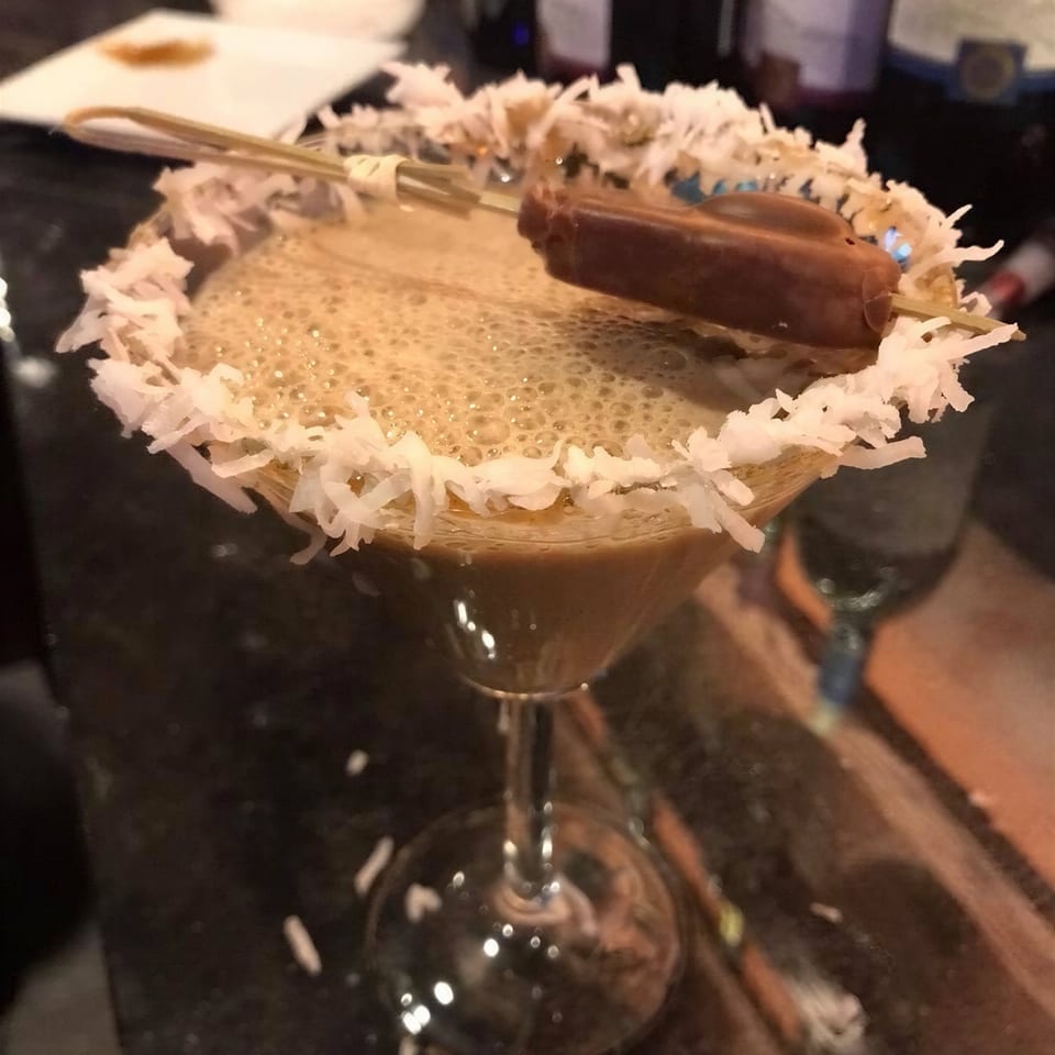Close-up of a cocktail glass rimmed with shredded coconut and garnished with a chocolate-covered treat, with bottles in the background.