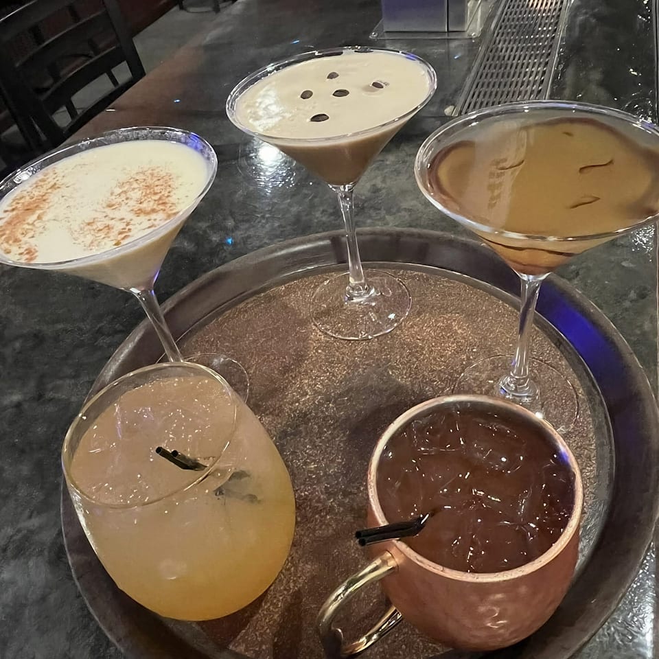 A tray holds five assorted cocktails, including creamy martinis, a pale citrus drink, and a dark drink, on a restaurant bar.