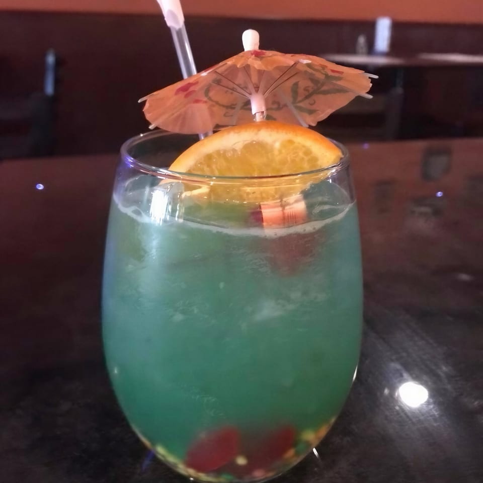 Blue-green cocktail in a stemless glass garnished with a lemon wedge, paper umbrella, and straw on a dark tabletop.