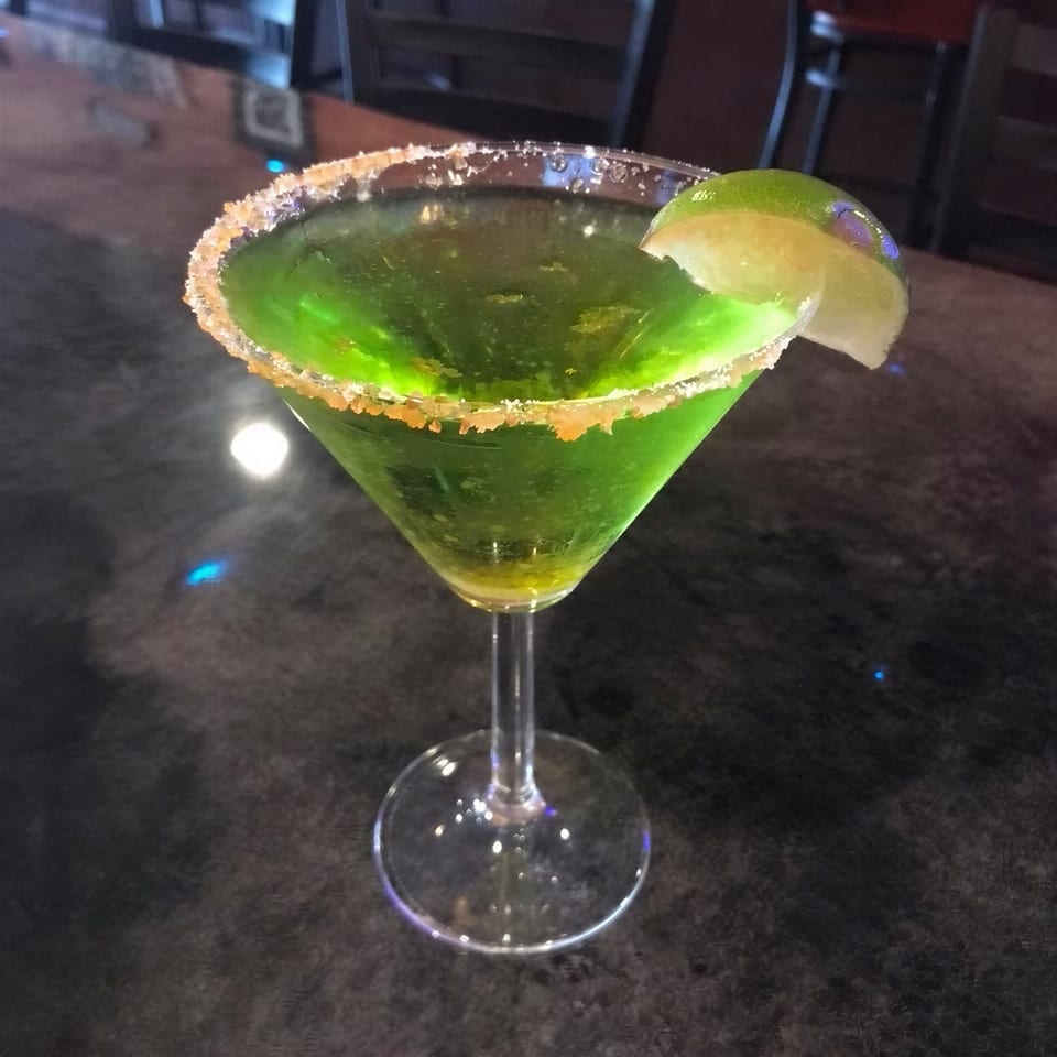 Bright green cocktail in a stemmed glass with a sugared rim and lime garnish on a restaurant table.